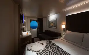 Interieur van de Deluxe Buitenhut OR op de World Atlantic met een comfortabel bed, zithoek en een karakteristiek rond raam met uitzicht op zee.